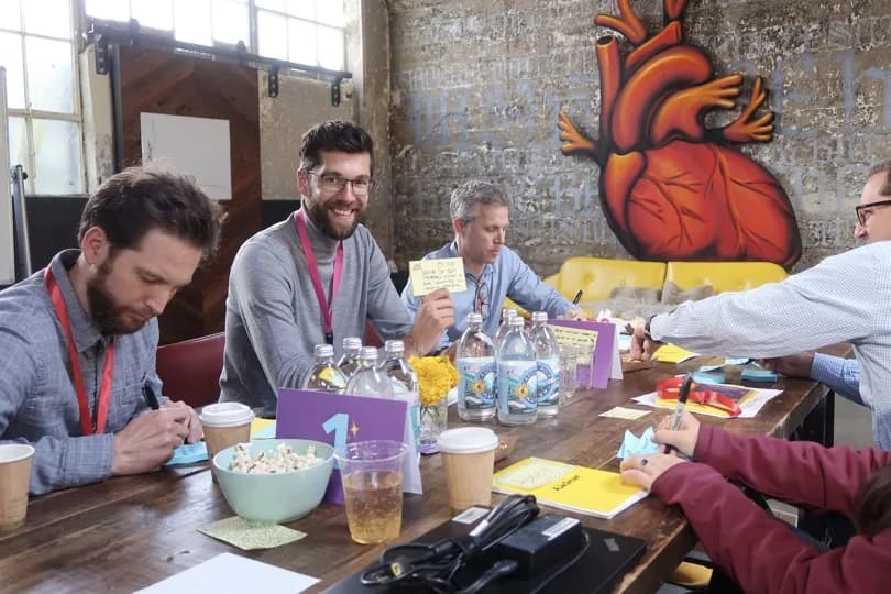 A workshop in progress: five people around a long table covered in sticky notes, markers and coffee cups, in a brick-walled room with a large painted anatomical heart on the wall.