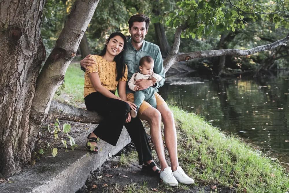 Danny, Jessie and baby Noa sitting together on a tree trunk by a river in a park.